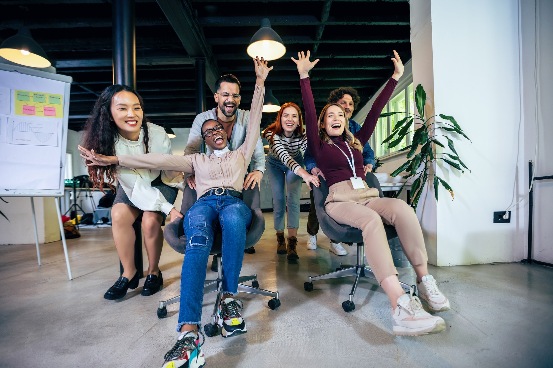 Young cheerful businesspeople in smart casual wear having fun while racing on office chairs and smiling